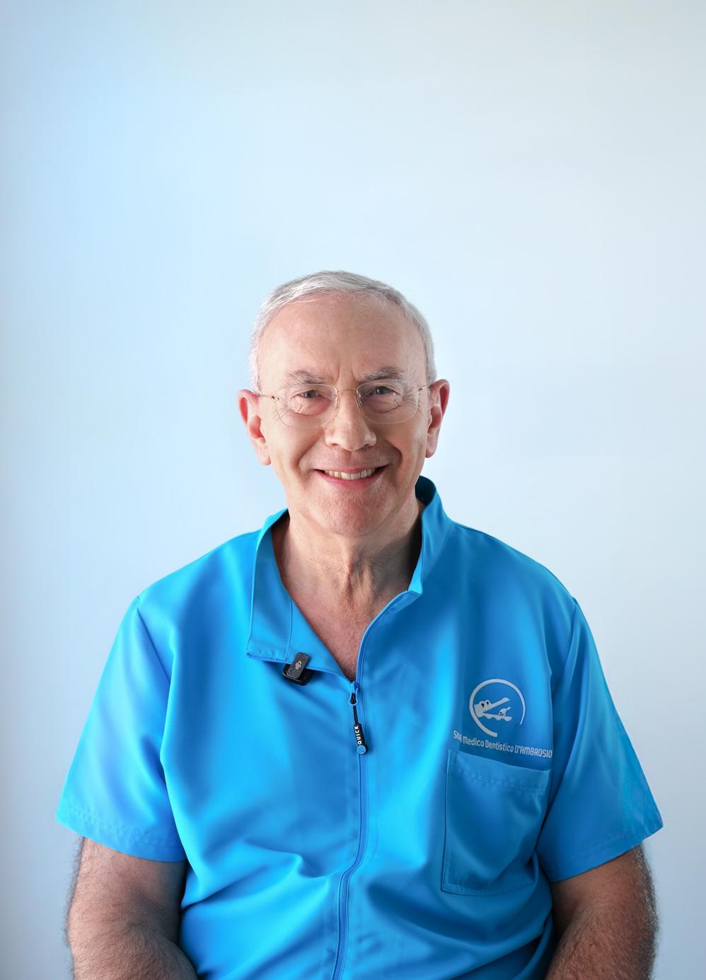 Ritratto di un medico odontoiatra in camice azzurro, seduto davanti a uno sfondo chiaro e sorridente verso la fotocamera.