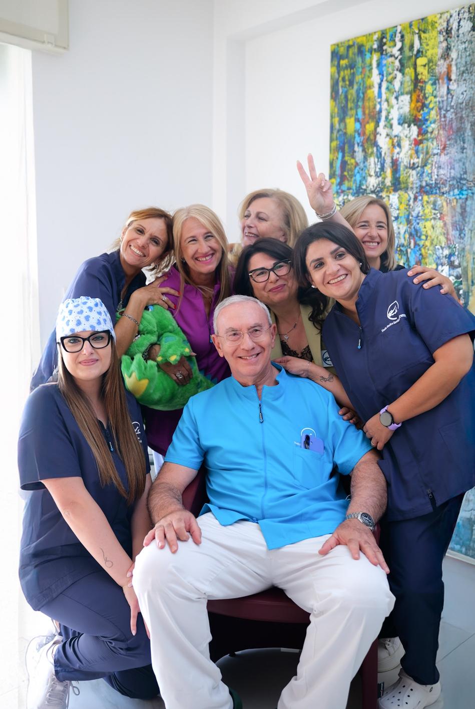 Gruppo di professionisti dello Studio Dentistico D’Ambrosio che posa sorridente in uno spazio luminoso; al centro un medico in camice azzurro, circondato da colleghe in divisa blu e viola.