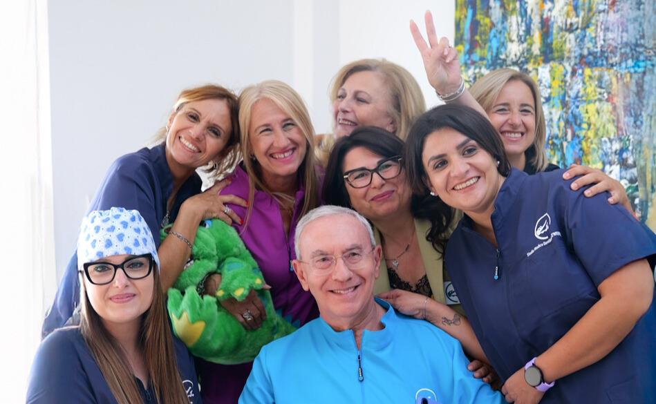 Gruppo di professionisti dello Studio Dentistico D’Ambrosio che posa sorridente in uno spazio luminoso; al centro un medico in camice azzurro, circondato da colleghe in divisa blu e viola.